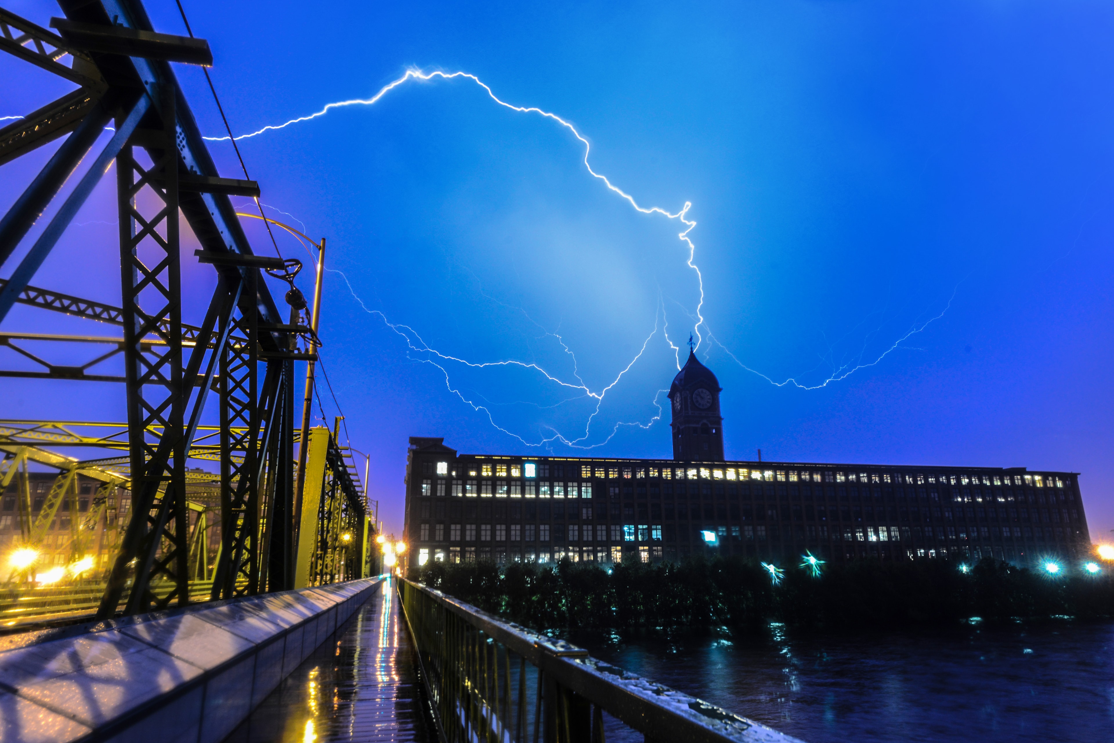 Lightning over a mill building and clock tower at night, photographed from a bridge by Scott Proposki